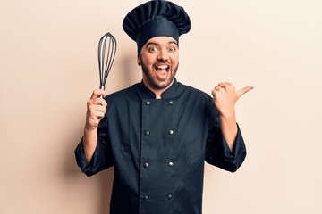 Fototapeta premium Young hispanic man wearing cooker uniform holding whisk pointing thumb up to the side smiling happy with open mouth