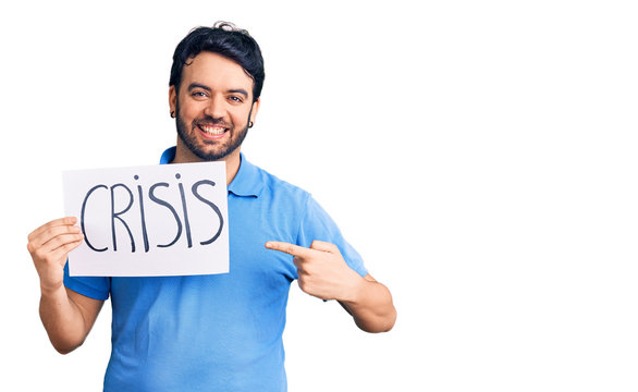 Young hispanic man holding crisis message paper smiling happy pointing with hand and finger