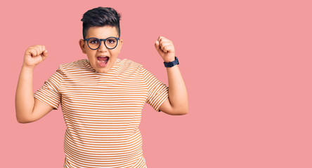 Little boy kid wearing casual clothes and glasses angry and mad raising fists frustrated and...