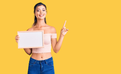 Young beautiful woman holding empty white chalkboard surprised with an idea or question pointing finger with happy face, number one