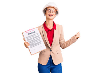 Young beautiful woman wearing architect hardhat holding clipboard with contract document screaming proud, celebrating victory and success very excited with raised arms