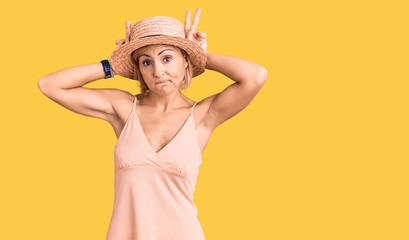 Young blonde woman wearing summer hat doing bunny ears gesture with hands palms looking cynical and skeptical. easter rabbit concept.
