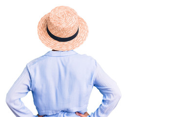 Young blonde woman wearing summer hat standing backwards looking away with arms on body