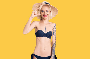 Young blonde woman with tattoo wearing bikini and summer hat smiling and confident gesturing with hand doing small size sign with fingers looking and the camera. measure concept.