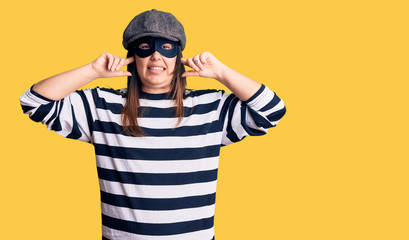 Young beautiful brunette woman wearing burglar mask covering ears with fingers with annoyed expression for the noise of loud music. deaf concept.