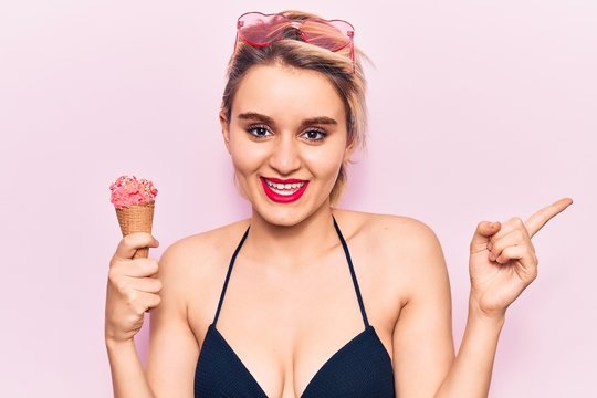 Young beautiful blonde woman wearing bikini holding ice cream smiling happy pointing with hand and finger to the side