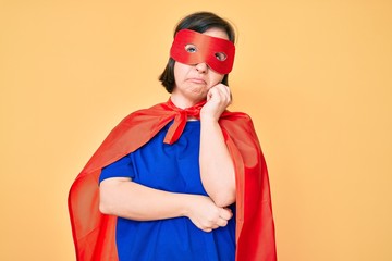 Brunette woman with down syndrome wearing super hero costume serious face thinking about question with hand on chin, thoughtful about confusing idea