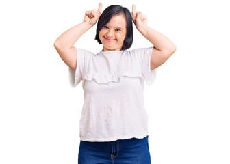 Brunette woman with down syndrome wearing casual white tshirt doing funny gesture with finger over head as bull horns