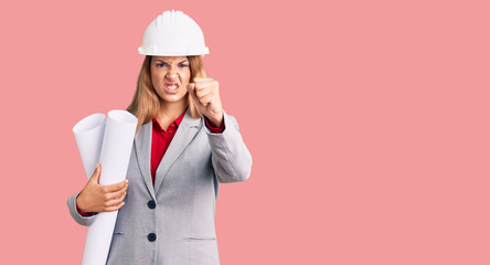 Beautiful young woman wearing architect hardhat holding build project annoyed and frustrated shouting with anger, yelling crazy with anger and hand raised