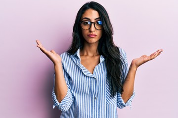 Beautiful hispanic woman wearing casual clothes and glasses clueless and confused with open arms,...