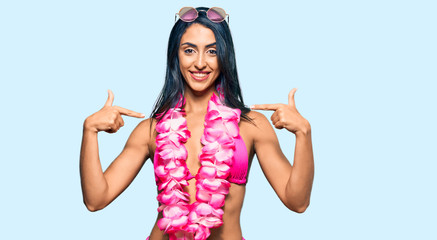 Beautiful hispanic woman wearing bikini and hawaiian lei looking confident with smile on face,...