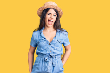 Brunette teenager girl wearing summer hat winking looking at the camera with sexy expression,...