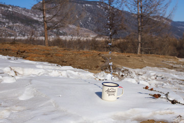 Drops of clean drinking water from Lake Baikal are splashed from a metal aluminum mug on the snow on a picturesque mountain background on a clear winter day among coniferous trees.