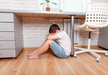 Preteen boy sitting under desk, covering his face with hands at home