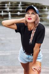 Young blonde with a braid in shorts and a cap. Beautiful smiling woman on the street of the summer city. Vertical.