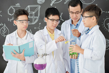 Chemistry teacher controlling school students mixing colorful liquids and taking notes in copybook