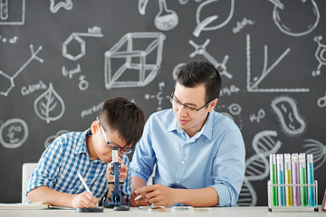 Vietnamese chemistry tutor explaining difficult topic to student and asking him to look at microscope and write dow his observations