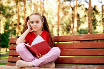 Obraz premium Little Girl Rest On Park Bench And Talk By Phone. Concept Of Speaking By Modern Gadgets And Fast Internet In Park.
