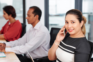 Portrait of happy Indian female entrepreneur talking on phone with business partner when sitting at office desk next to colleagues