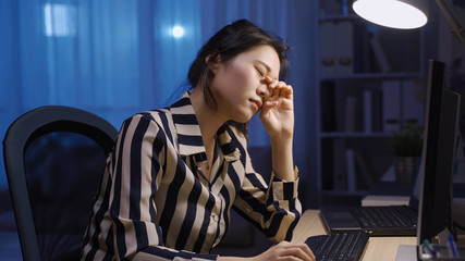 asian businesswoman is opening eyes wide trying to concentrate while working overtime at midnight. taiwanese lady propping head is rubbing sore eyes while using desktop. healthcare and work from home - obrazy, fototapety, plakaty