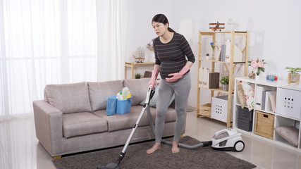 full length asian female in her third trimester feeling sudden painful contraction while cleaning floor with a hoover at home. stroking belly with discomfort facial expression. genuine lifestyle