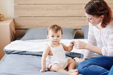 Young mother playing with her cute serious little daughter sitting on bed