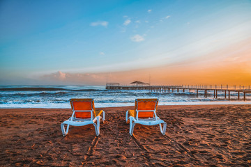 Sunbed on the sand beach and view to water of sea, waves with white foam, pierce and sky with clouds in a nice evening during sunset. The concept of a holiday on the sea