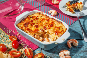 Traditional italian lasagna with vegetables, shrimps and cheese in a blue casserole dish and and part of dish on plate with overturned glass of wine on a pink and blue background. 