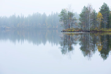 Fototapeta na wymiar Early foggy morning by the lake. Trees in autumn colors are reflected on the surface of the lake, distant trees are wrapped in the fog