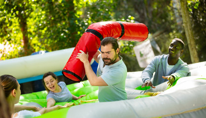 Adults having fun on inflatable amusement playground. Expressive bearded guy fighting off his friends with inflatable log while they trying to filch toy chickens