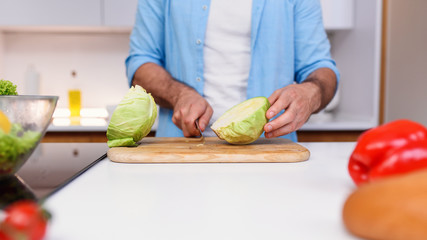 Fototapeta na wymiar Caucasian man in blue shirt cuts the cabbage with a knife on a wooden board at cozy home kitchen.