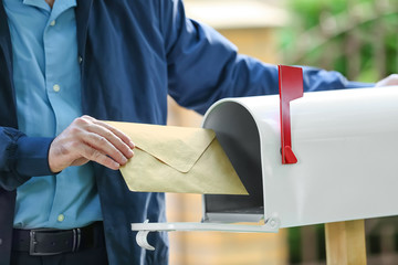 Handsome young postman putting letter in mail box outdoors - Powered by Adobe