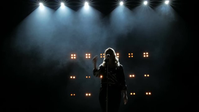 Silhouette of a girl comes to the microphone on the stage against a background of bright yellow lights