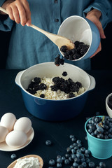Woman with wooden spoon put blackberry into bowl with cottage cheese. Cottage cheese casserole cooking.