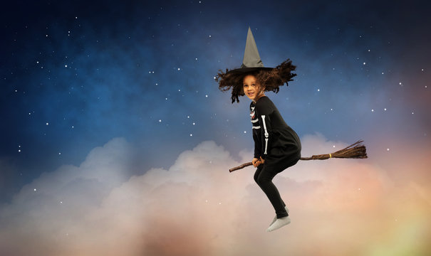 halloween, holiday and childhood concept - smiling african american girl in black costume dress and witch hat with broom over starry night sky and clouds on background