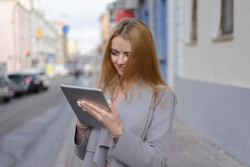 girl with red hair in coat looks something in a tablet on a city