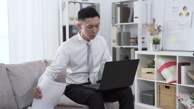 asian guy seated on sofa is preparing financial statement with notebook and document. man working from home is contrasting information on paper and entering number on pc