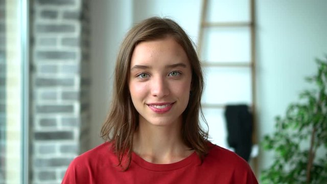 Portrait of beautiful young woman posing in front of camera while standing in light interior avki. Face view of American girl looks forward with toothy smile and poses at room with daylight. One