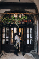 Fototapeta na wymiar A young caucasian girl in an autumn dark coat and a wide scarf with black gloves stands in the doorway and looks into the distance. Old door with glass. Lviv, Italian courtyard, Ukraine.