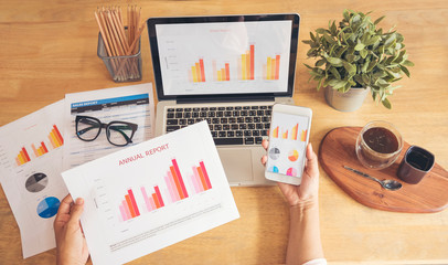 Woman working at home office desk using laptop business financial document chart and graph on wooden table with coffee cup. Woman freelance reading business graphs sale report on business office desk