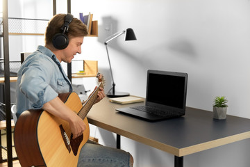 leisure, music and people concept - young man or musician in headphones with laptop computer playing guitar sitting at table at home