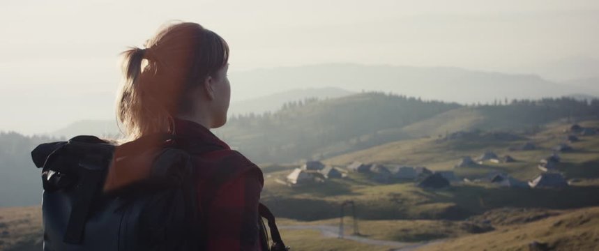 Woman hiking look at scenic view of Velika Planina in Europe Alps mountain cottage village on a hike in fall or autumn morning over shoulder close up slow motion flare