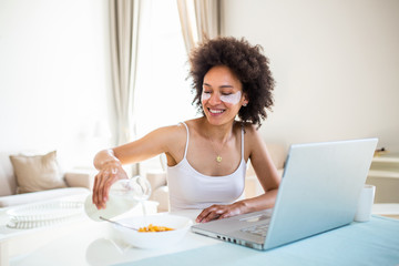 Close up smiling young mixed race woman pours corn flakes in plate with milk. The girl has a healthy breakfast on stylish cozy home at the morning while checking her email on laptop.