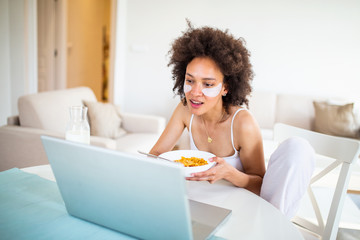 Obraz na płótnie Canvas Charming young african american woman sitting in the morning at the table browsing notebook, freelancer have breakfast eat cereal corn flakes, stay home distance remote work quarantine indoors