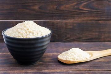 A black ceramic bowl and wooden spoon filled with uncooked white rice. Light grains of cereals on a dark brown background. Organic natural healthy food. An ingredient for traditional Asian cuisine.
