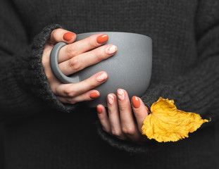 Woman holds a cup of hot tea. Grey knitted sweater, a mug of warm drink. Female hands with trendy autumn marble manicure in orange color