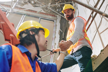 Serious bearded builder helping coworker to climb up the high stairs in building under construction