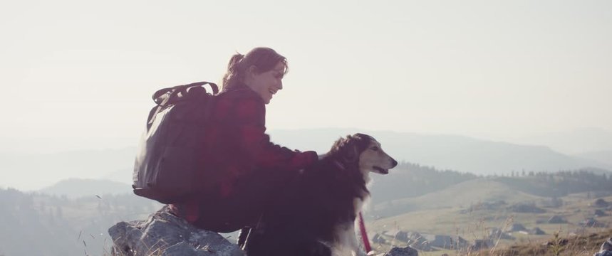 Woman on a hike sit on top view pet her dog drink from water bottle smile and happiness in the morning light hiking in fall or autumn at Velika Planina in Europe Alps handheld with sun flare