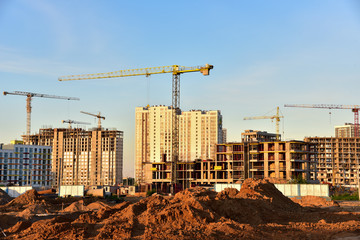 Fototapeta premium Tower cranes working at construction site on blue sky background. Construction process of the new modern residential buildings. Construction roads and streets in city. Soft focus