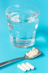 A glass of water and three capsules and spoon with collagen powder on blue background.